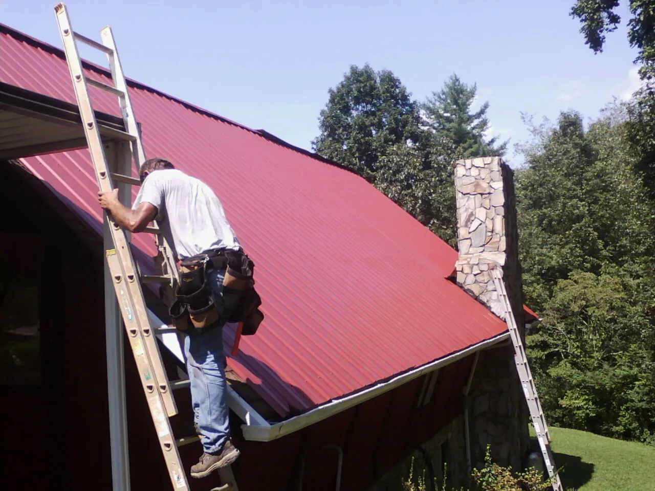 Roofer on a ladder beside a red metal roof and stone chimney — Soffit & Fascia Repair by Roofing Brothers in Fryeburg
